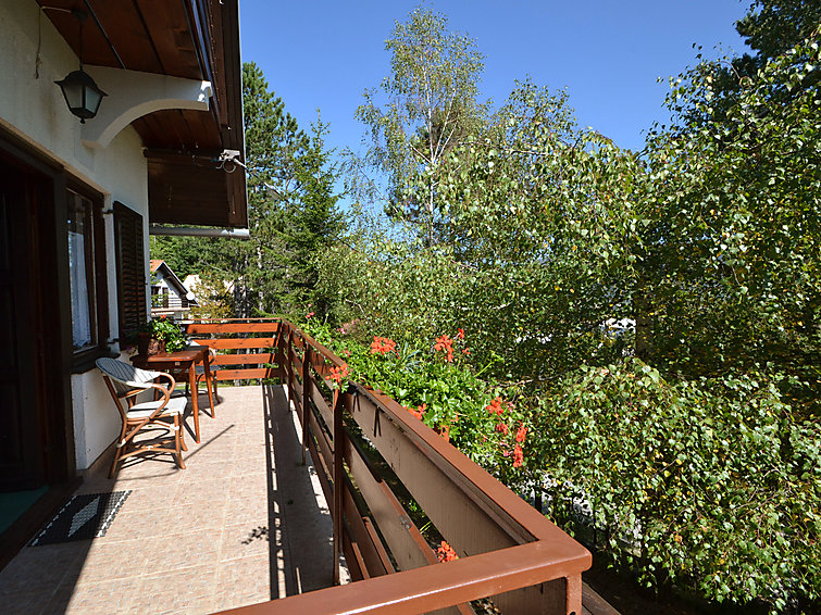 Balkon kuće s pletenom foteljom i stolićem, narančaste geranije na drvenoj ogradi, breze i plavo nebo iza